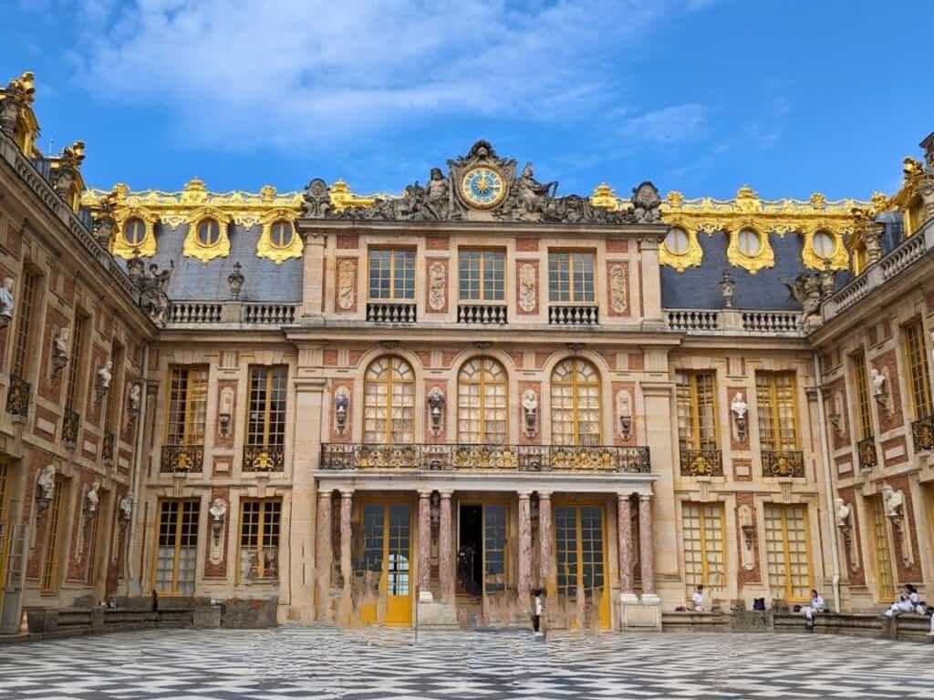 The ornate facade of the Palace of Versailles features gold details, arched windows, statues, and a patterned courtyard under a blue sky—a breathtaking sight on any Versailles tour.