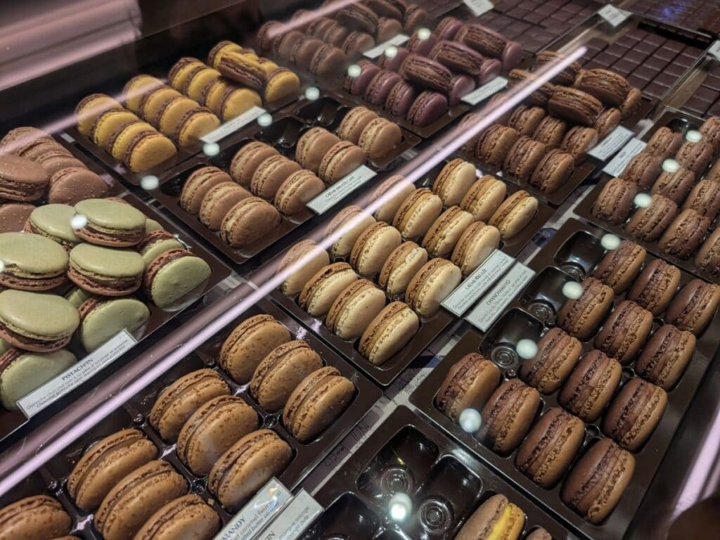 Rows of assorted macarons and chocolates are displayed in a glass bakery case, organized by flavor and color on brown trays—an inviting scene for anyone ready to eat your way through Paris on the ultimate food tour.