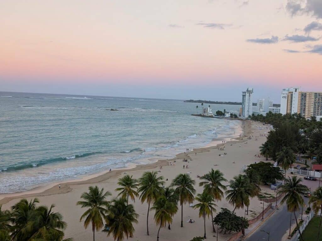 A wide sandy beach in San Juan, Puerto Rico, with palm trees, gentle waves, and scattered people at sunset, high-rise buildings lining the shoreline—the perfect spot to spend 48 hours unwinding by the sea.