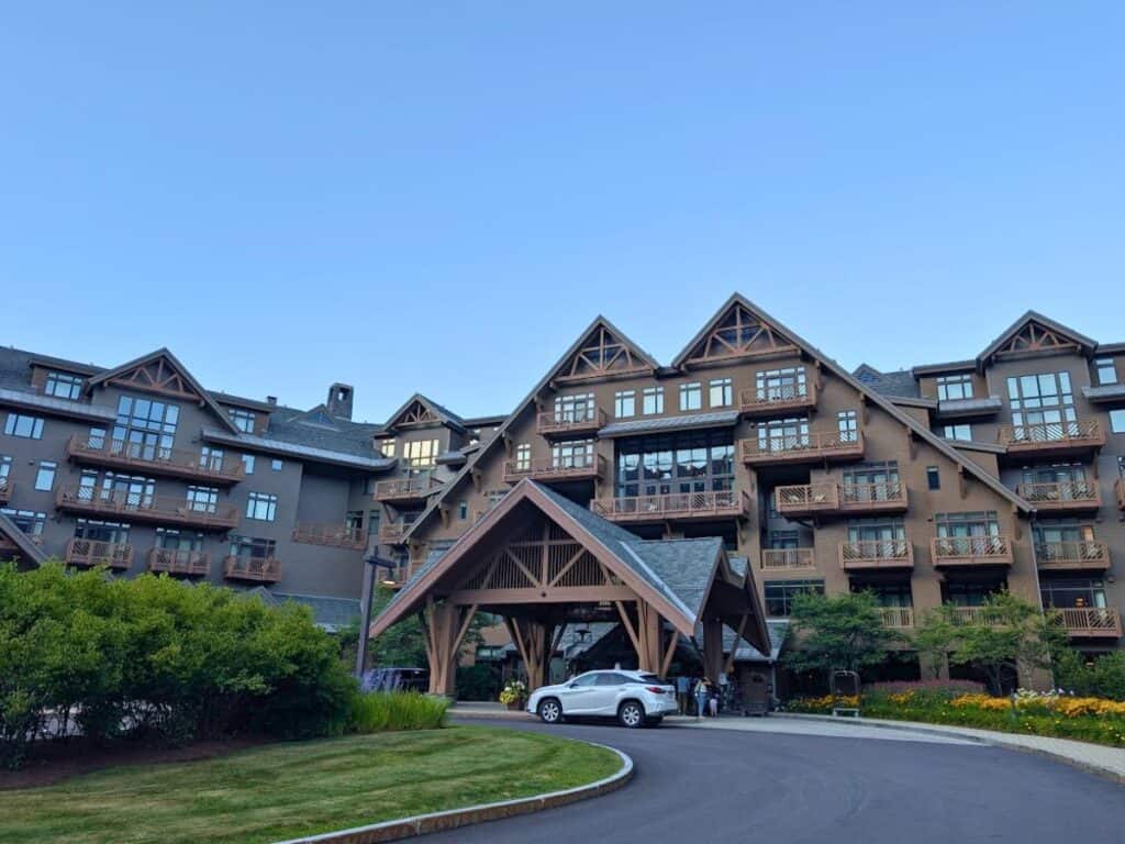 Large, multi-story lodge-style building with wooden balconies and a covered driveway entrance; a white car is parked at the front of the Lodge at Spruce Peak, surrounded by lush landscaping and greenery.