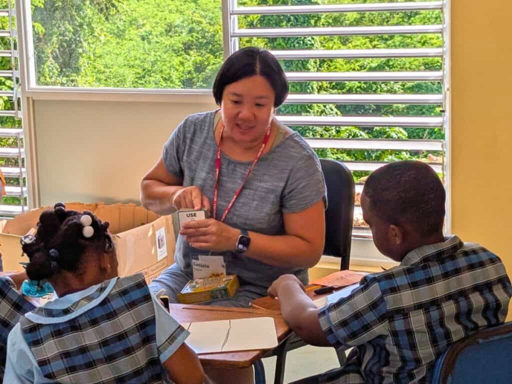 A woman sits at a table in Jamaica, volunteering as she helps two children in matching plaid uniforms with an activity. Supplies are spread across the table.
