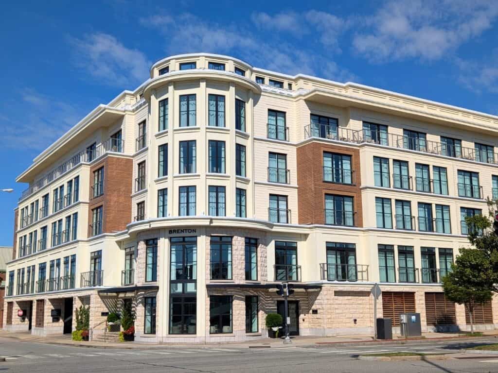 A modern four-story building with large windows, balconies, and a rounded corner entrance labeled "The Brenton" offers an ideal stay in Newport on a sunny day.