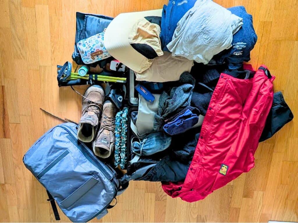 Assorted hiking and camping gear, including boots, backpack, jacket, hat, clothing, water bottle, gloves, and various small outdoor essentials laid out on a wooden floor—perfect for any Patagonia packing list.