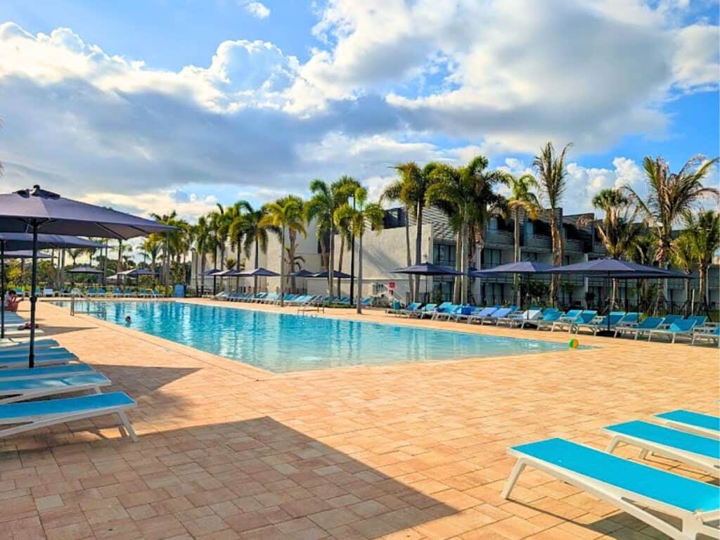 All-inclusive outdoor swimming pool surrounded by blue lounge chairs and umbrellas, with palm trees and a modern hotel building in the background under a partly cloudy sky.
