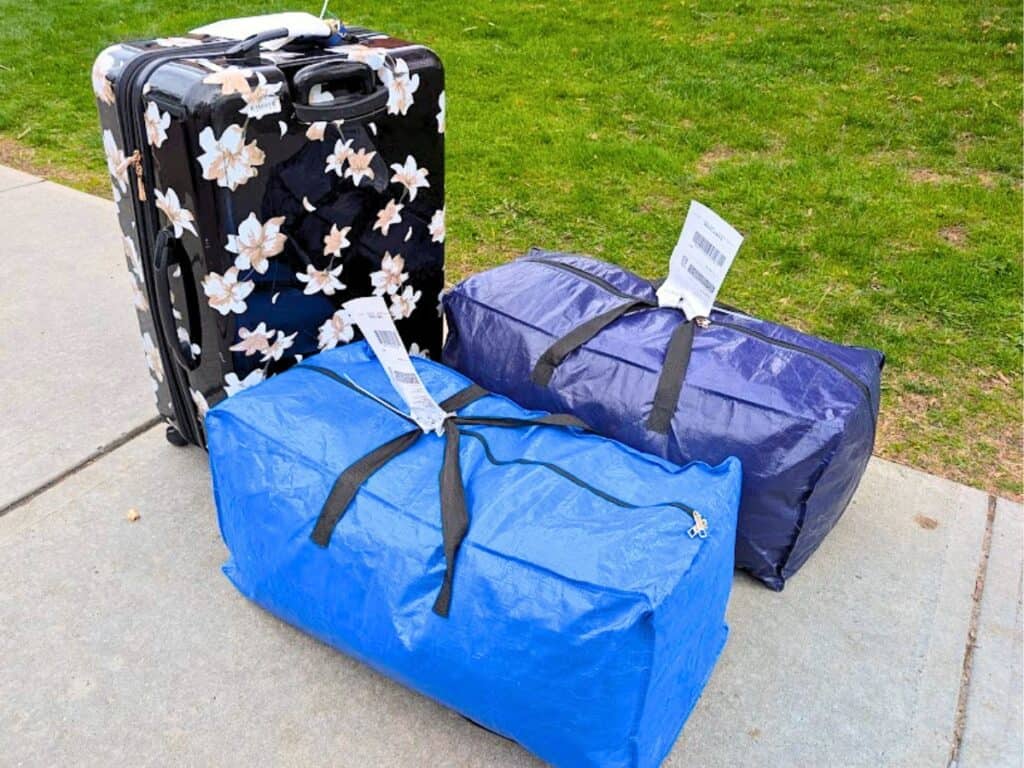 Three labeled pieces of luggage—one floral-patterned suitcase, one blue duffel bag, and one dark purple duffel bag—are on a sidewalk next to grass, ready for airport luggage shipping.