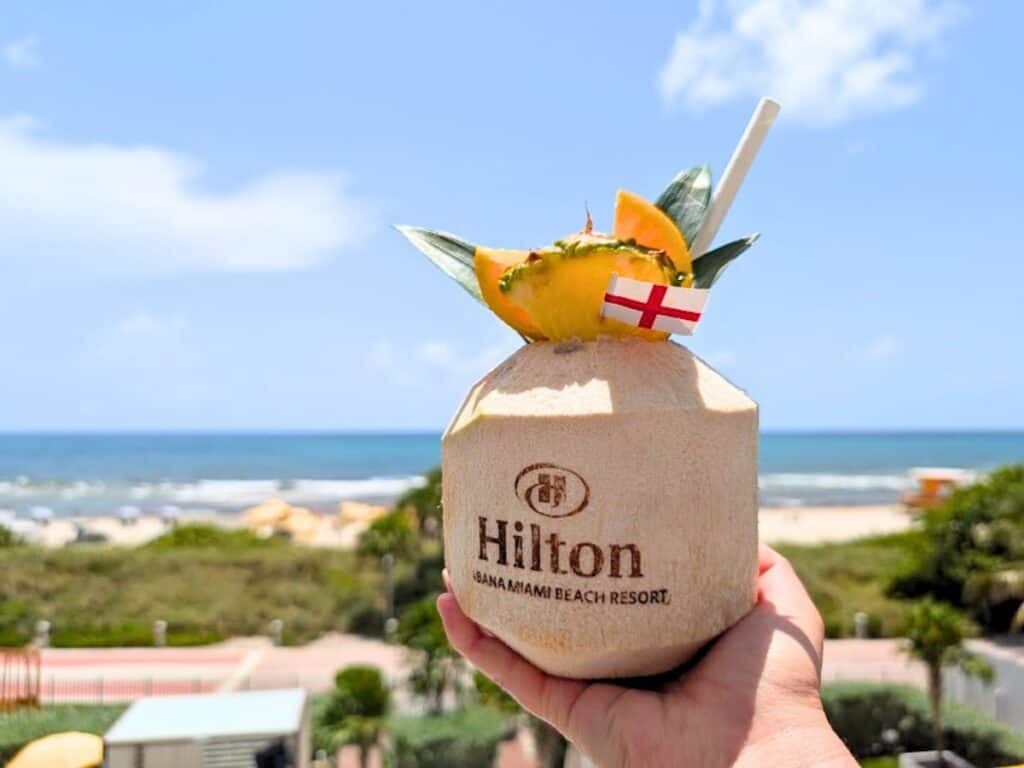 A hand holds a Hilton-branded coconut drink with tropical garnish and a small flag, set against the backdrop of a stunning beach and ocean under a clear blue sky—perfect for capturing that unique Hilton Cabana Miami Beach Resort review moment.
