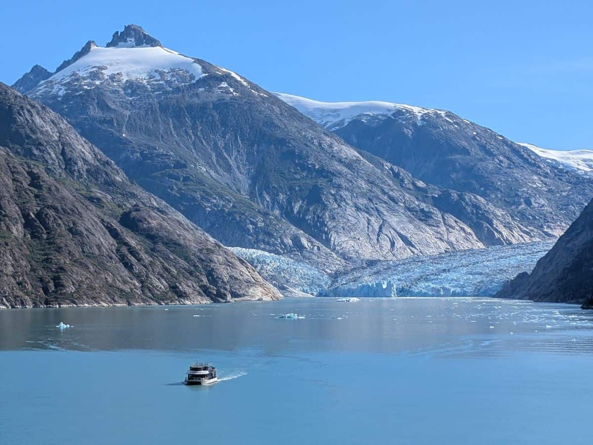 A boat sails on a calm blue fjord toward a glacier, surrounded by tall, snow-capped mountains under a clear sky.
